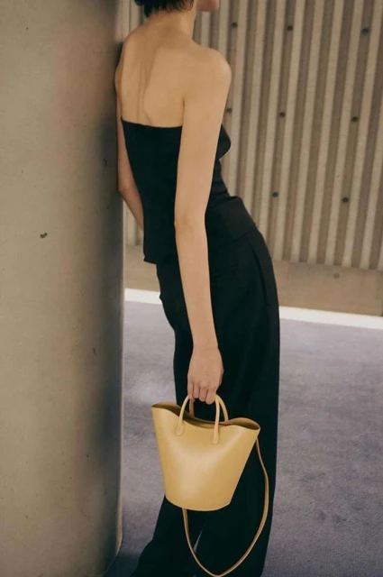 A woman in a black strapless top and trousers poses against a grey wall, holding a tan leather handbag with handles.