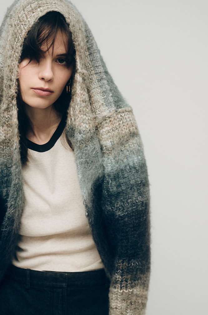 A young woman with dark hair wears a hooded, ombre knit cardigan over a black-trimmed t-shirt against a plain light background.
