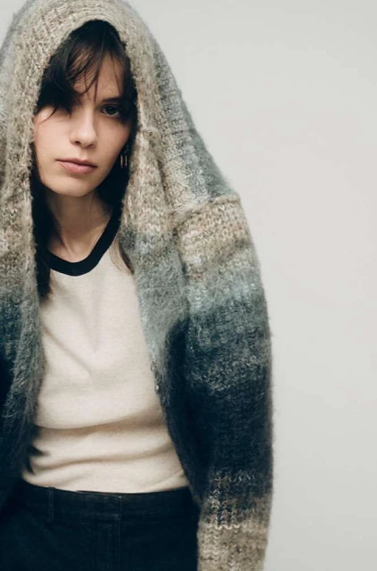 A young woman with dark hair wears a hooded, ombre knit cardigan over a black-trimmed t-shirt against a plain light background.