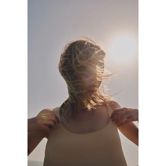Woman with blonde hair stands outdoors on a sunny day. She wears a tank top, and the bright sun is behind her. Wind blows her hair.