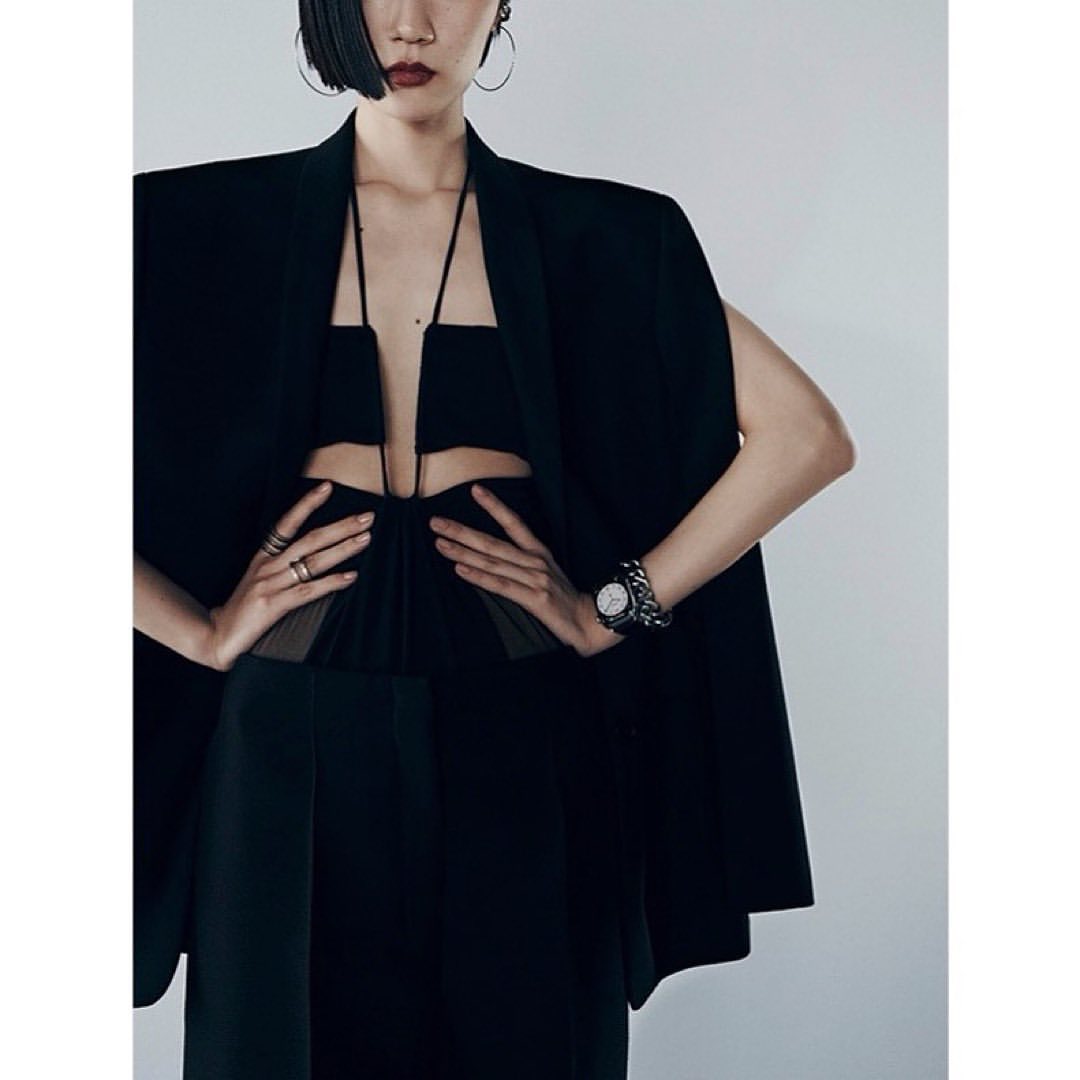 Elegant woman in a black dress with cutout details and a black cape, accessorized with hoop earrings, rings, and a watch. Studio setting.