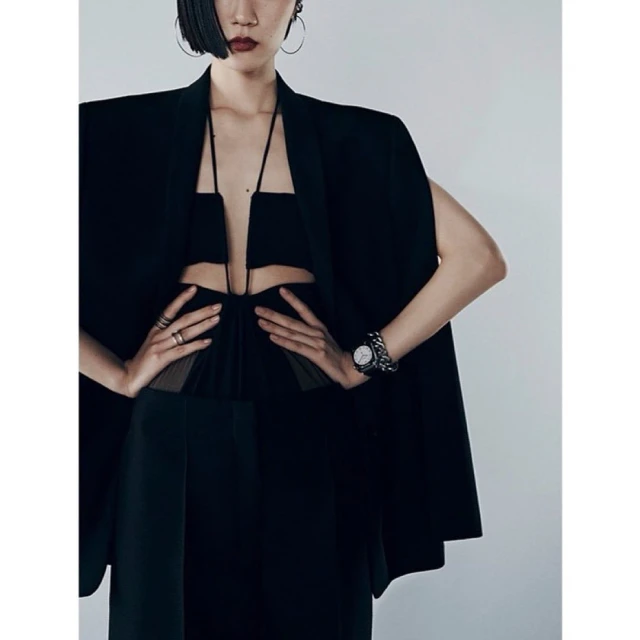 Elegant woman in a black dress with cutout details and a black cape, accessorized with hoop earrings, rings, and a watch. Studio setting.