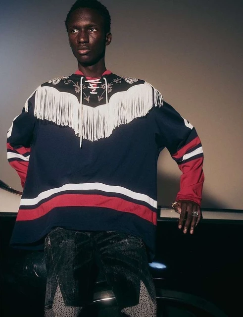 A person with dark skin and short hair poses in a dark blue shirt with fringed trim on the shoulders and chest in a studio.