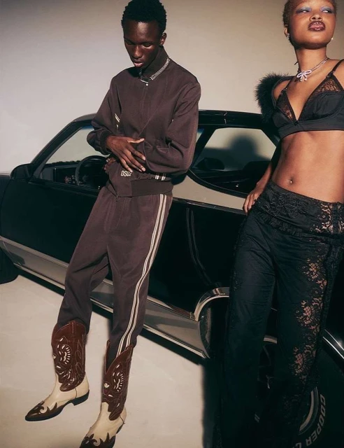 Two models pose in front of a black car, wearing fashionable streetwear outfits. The scene is simple and stylish with a neutral background.