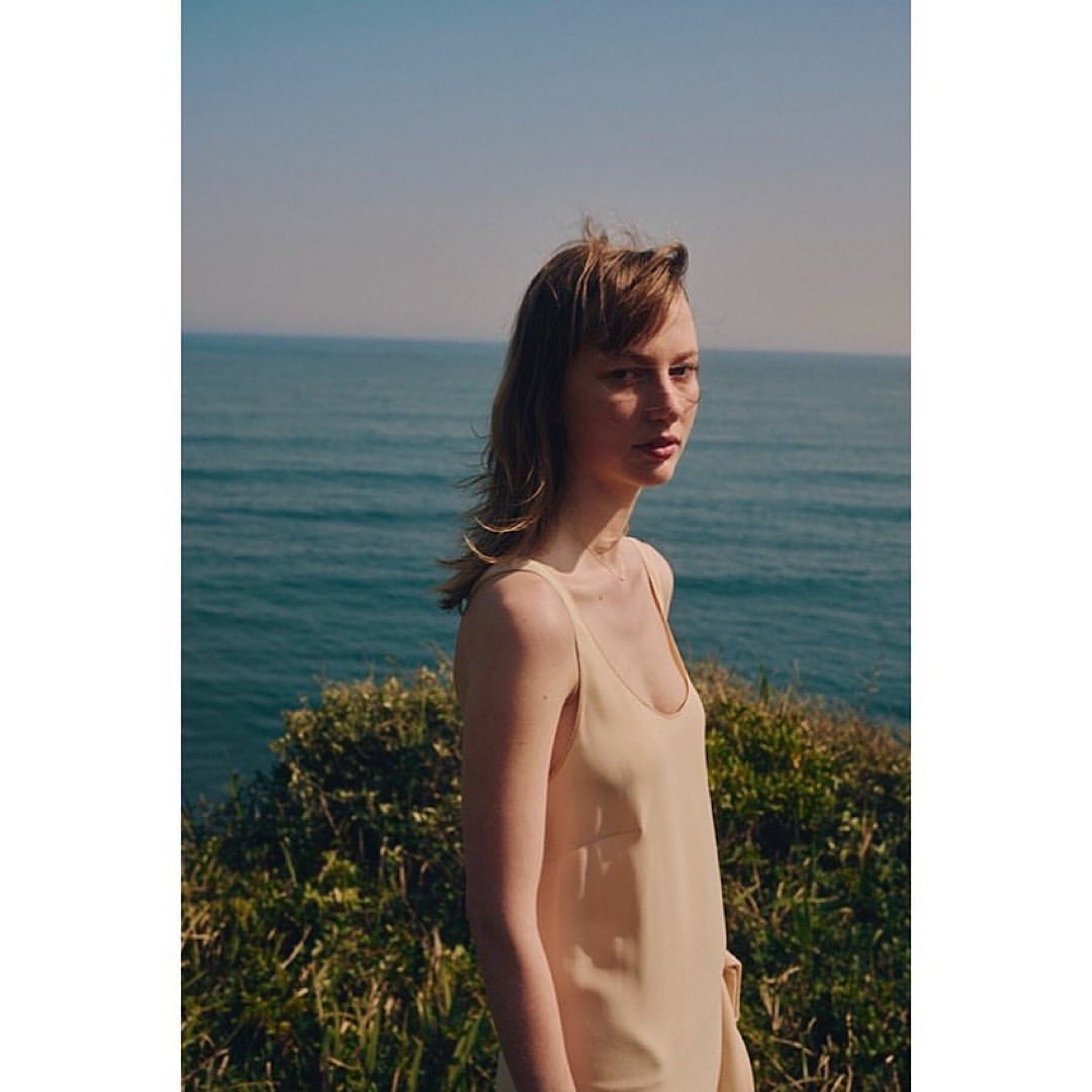 A woman in a cream slip dress standing outdoors with the ocean and green brush in the background.