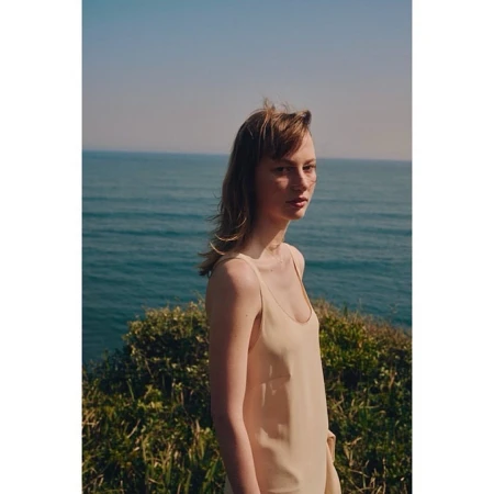 A woman in a cream slip dress standing outdoors with the ocean and green brush in the background.