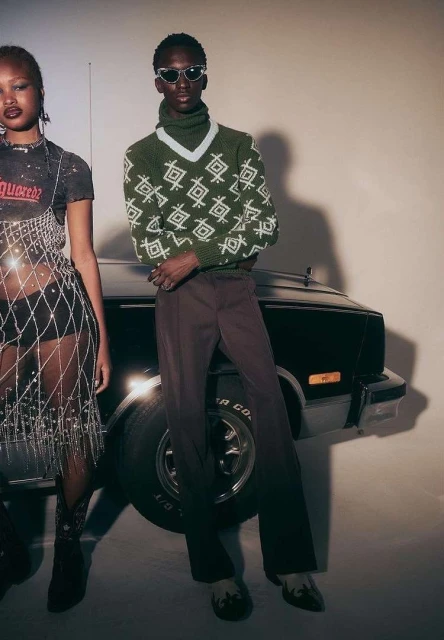 Two models pose in a studio, one in a mesh top and the other in a patterned sweater, standing in front of a vintage car.