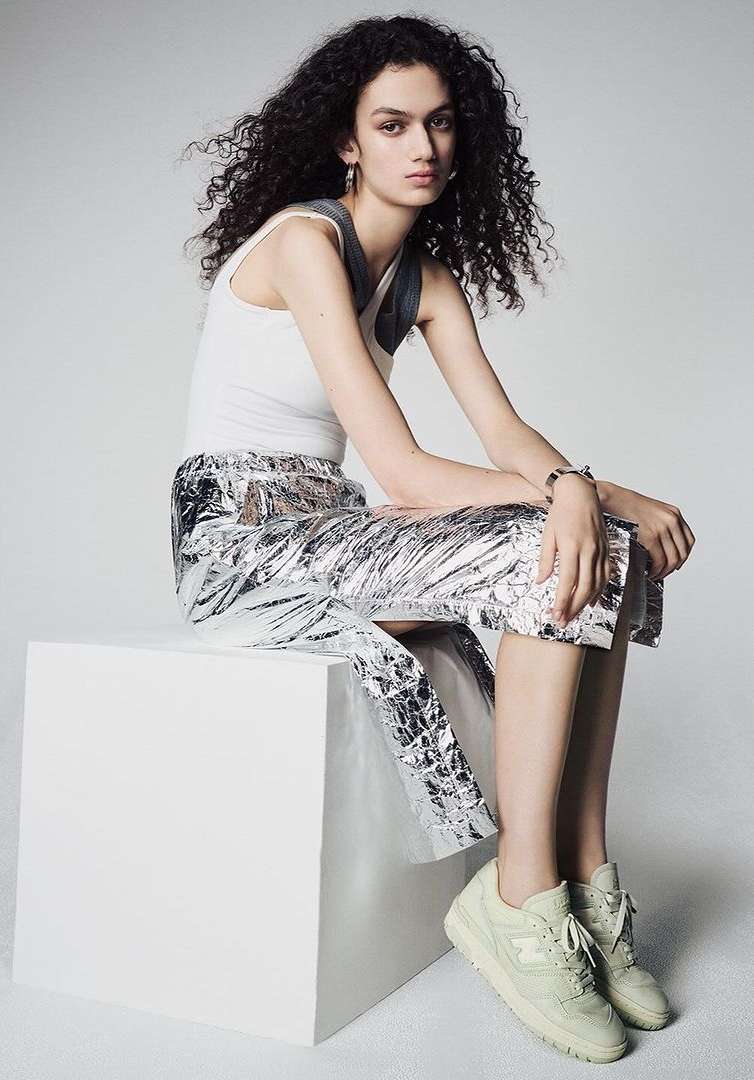 Young woman with curly hair posing seated on a white block wearing a white top and shiny silver pants and light-colored sneakers.