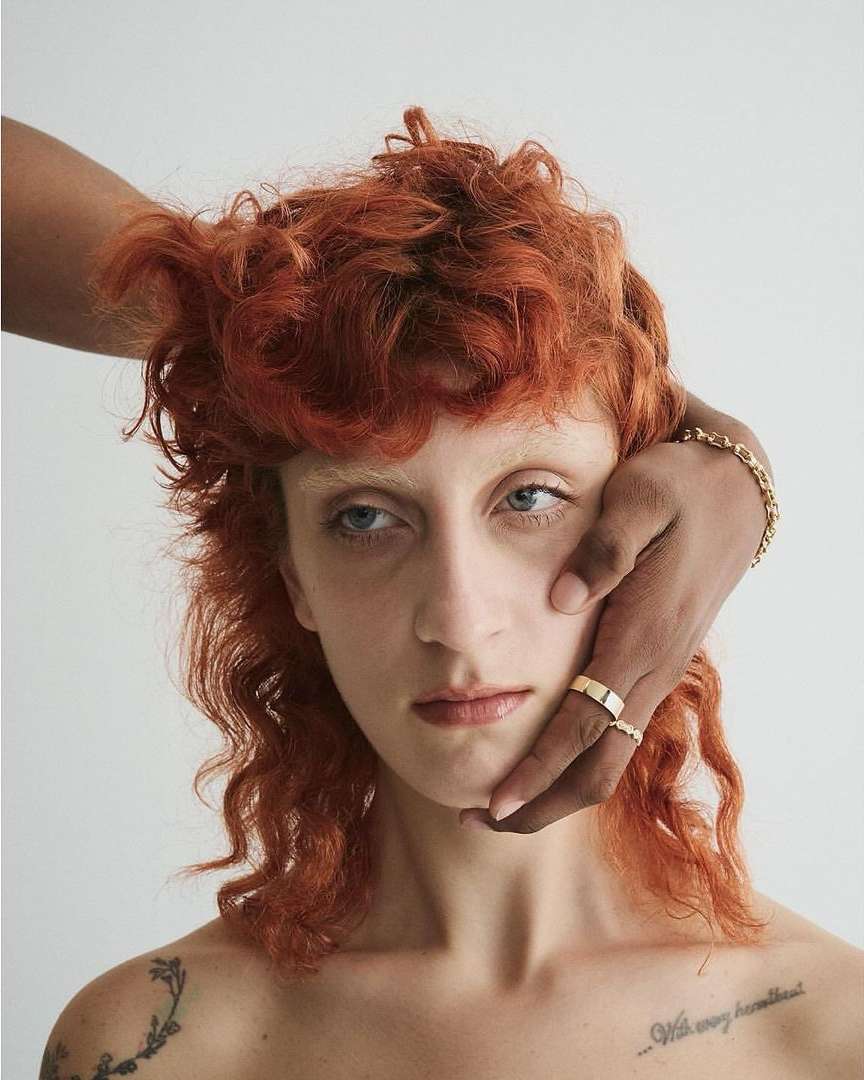 Close-up studio shot of a fair-skinned woman with curly red hair and hands of a dark-skinned person touching her face.