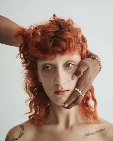 Close-up studio shot of a fair-skinned woman with curly red hair and hands of a dark-skinned person touching her face.
