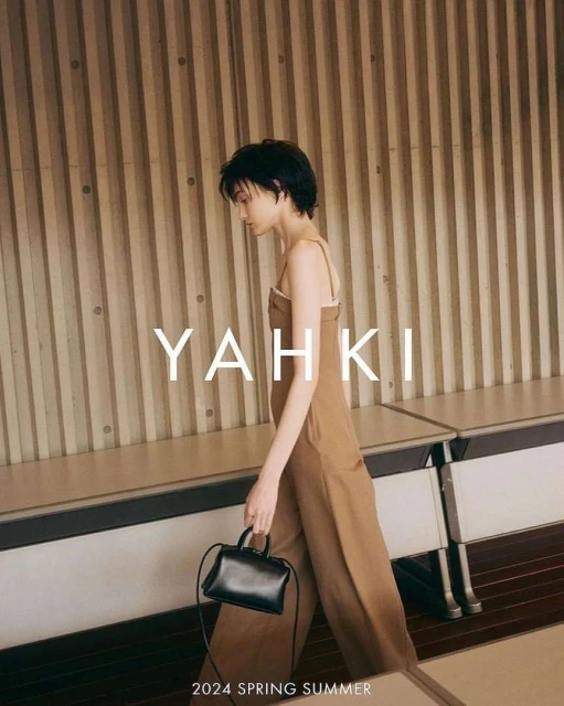 A woman models a brown jumpsuit and black purse, walking in front of a textured wall and tables for a fashion editorial shoot.