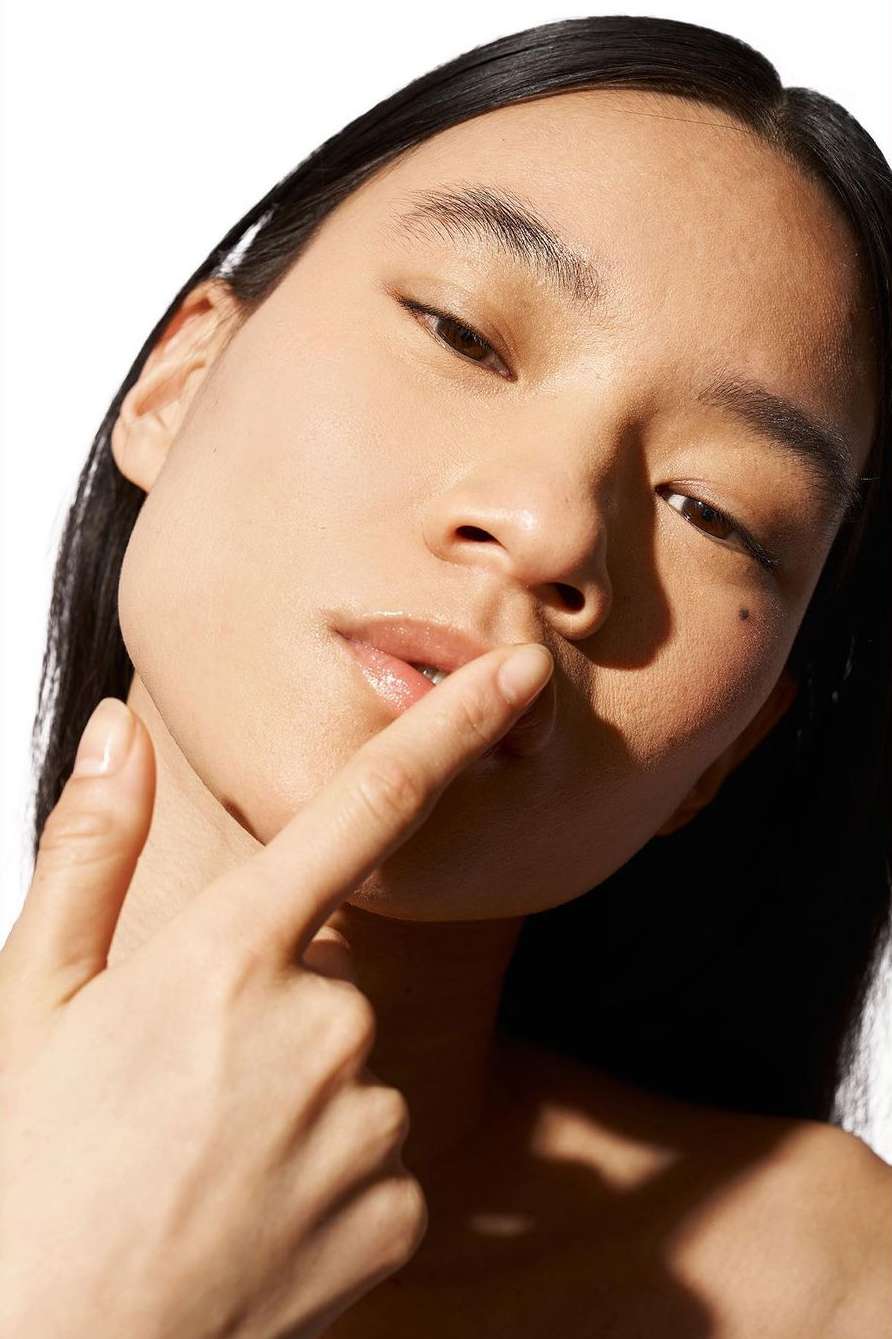 Close-up portrait of a woman with dark hair touching her lip, against a bright white background, showcasing natural beauty.