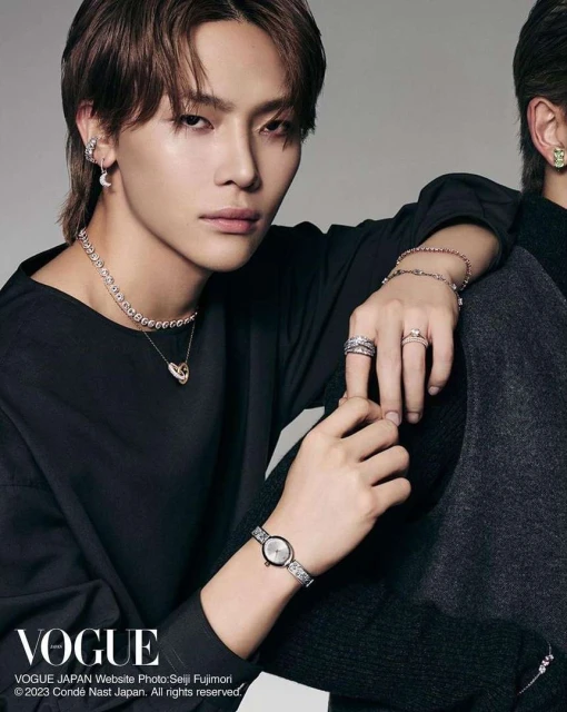 Asian male fashion model with dark hair, black top, jewelry (necklace, rings, watch, earrings) sitting next to another man indoors.