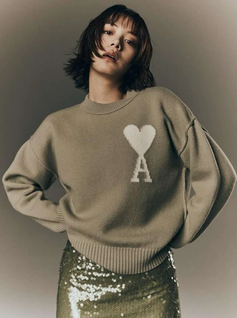 Brunette woman in a grey sweater with a heart logo and a shiny skirt posing indoors for a fashion shot in a studio.