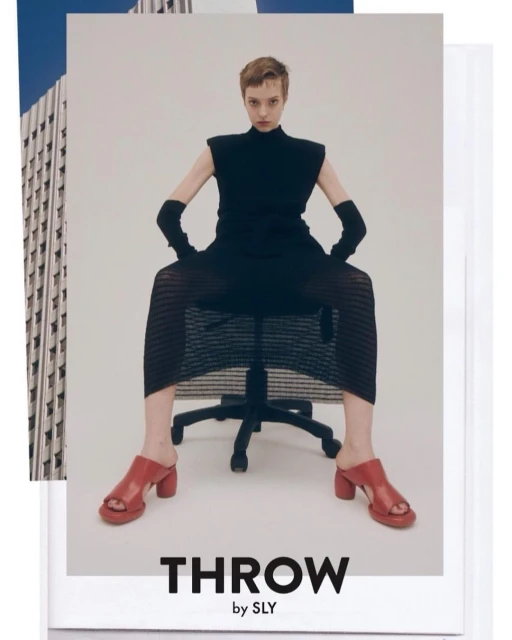 Fashion editorial shot of a woman in a black dress and red sandals, sitting on an office chair against a collage of a building.