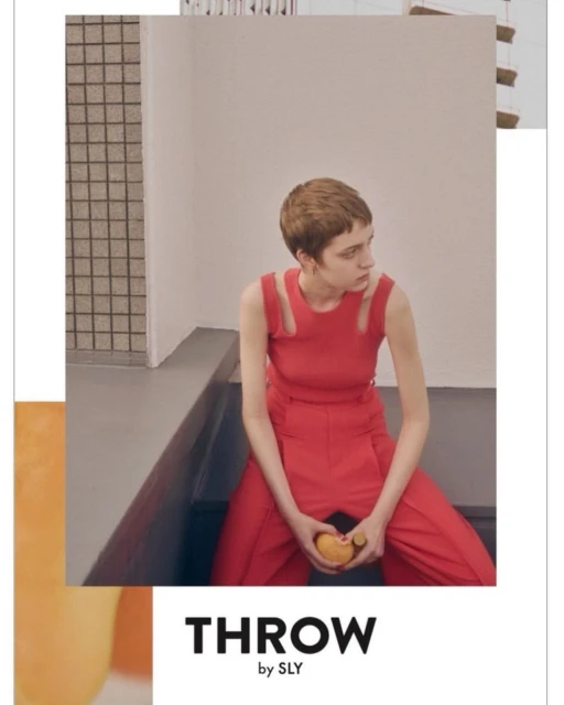 A woman with short hair poses in a red sleeveless top and matching pants while seated indoors, holding an orange, styled in a modern fashion.