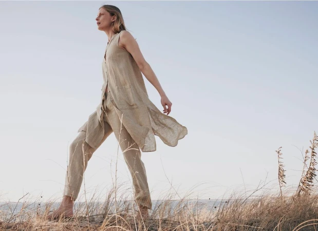 Woman walking barefoot in a beige linen sleeveless tunic and trousers through tall coastal grass against a clear sky