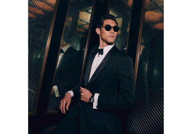 Man wearing a black tuxedo, bow tie, and round sunglasses in an elevator with mirrored walls.
