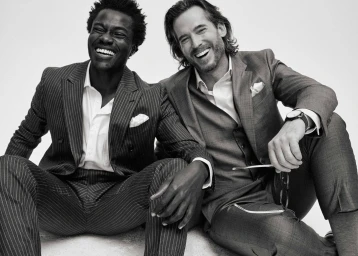 Two smiling men in tailored suits seated together against a plain white background in a black and white portrait.