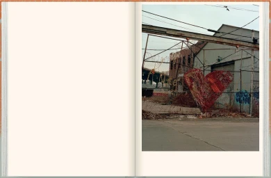Open book spread showing a blank cream page on the left and an urban street photograph of a red heart sculpture on a chain-link fence on the right.