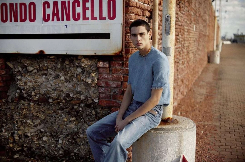 Young man in a blue knit shirt and jeans sits on a concrete post outdoors in front of a brick wall with a sign in natural lighting.