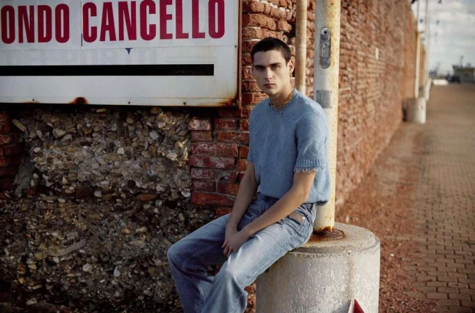 Young man in a blue knit shirt and jeans sits on a concrete post outdoors in front of a brick wall with a sign in natural lighting.