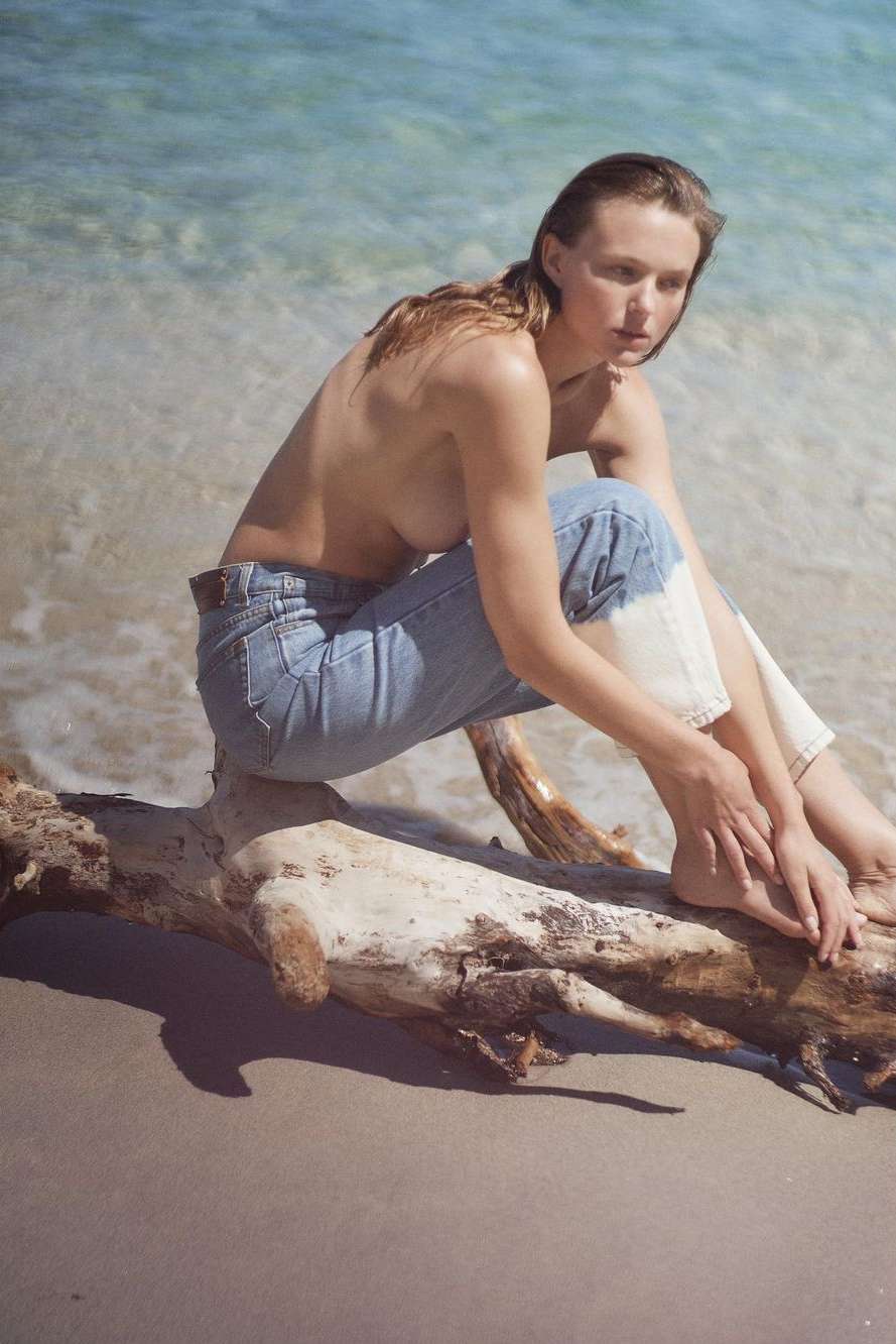 Woman in jeans sits on driftwood at the beach with turquoise water in the background in a sunny outdoor environment.