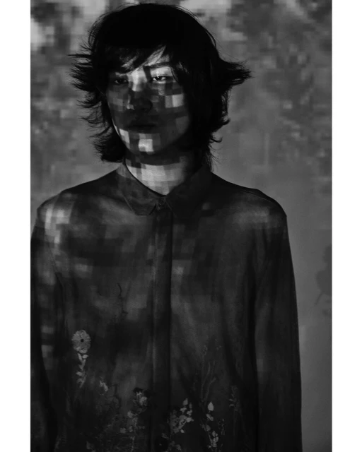 Monochrome portrait of a stylish person with dark hair, wearing a patterned button-down shirt, lit by pixelated light patterns indoors.
