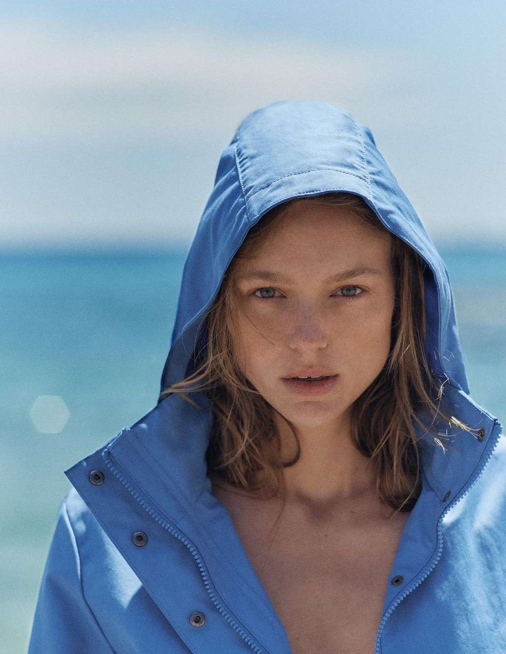 Portrait of a fair-skinned woman in a blue hooded jacket looking directly at the camera against a blurred background of the ocean and sky.