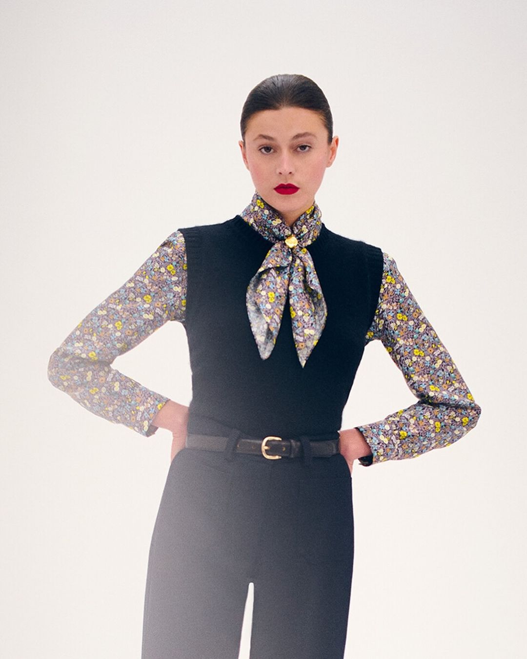 Elegant adult woman in a floral blouse, black sweater vest, and matching slacks poses against a clean, bright white backdrop.