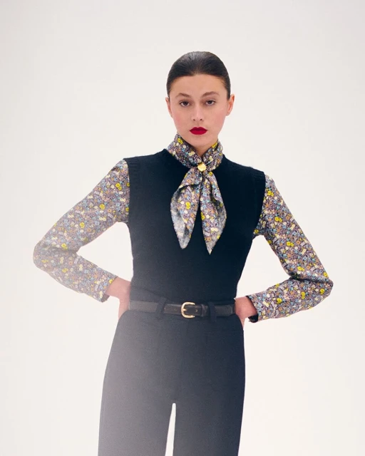 Elegant adult woman in a floral blouse, black sweater vest, and matching slacks poses against a clean, bright white backdrop.