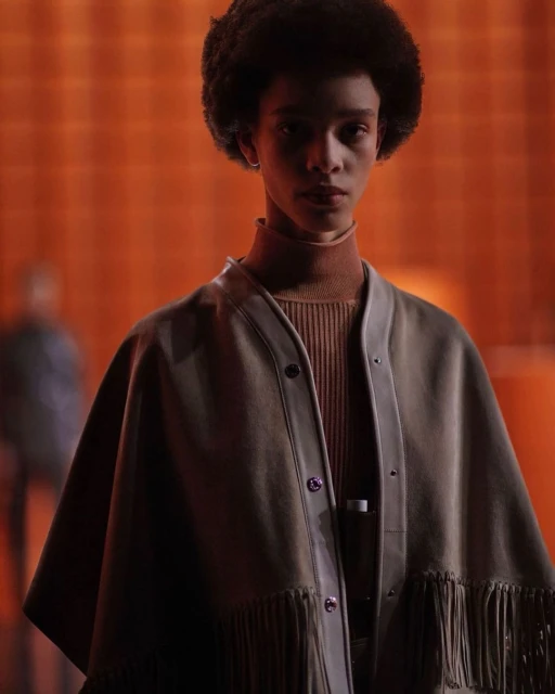 A person with afro hair wearing a tan cape with fringe and a brown turtleneck sweater, posing indoors against an orange background.