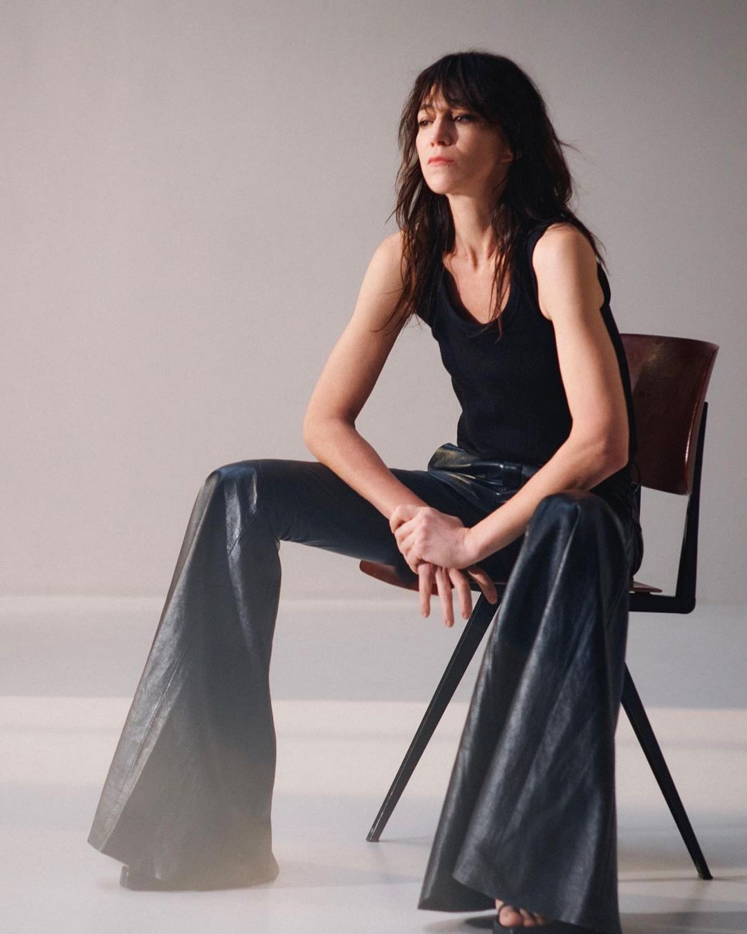 Portrait of a woman with dark hair in a black tank top and shiny black pants sitting on a chair in a brightly lit studio.