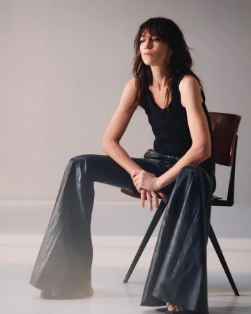 Portrait of a woman with dark hair in a black tank top and shiny black pants sitting on a chair in a brightly lit studio.