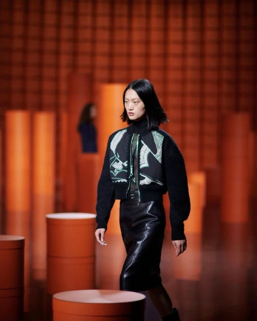 Runway fashion show featuring an Asian woman in a black bomber jacket and leather skirt, walking past orange cylinder props and background.