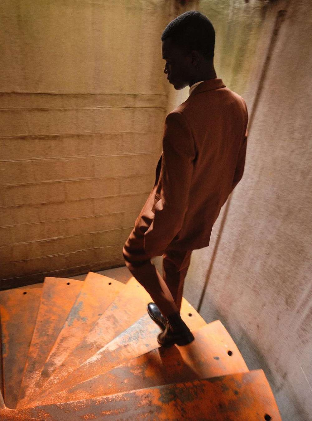 Man descends spiral staircase in suit. Angular light highlights rusty stairs and textures of the bare concrete walls.