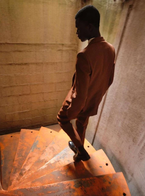Man descends spiral staircase in suit. Angular light highlights rusty stairs and textures of the bare concrete walls.