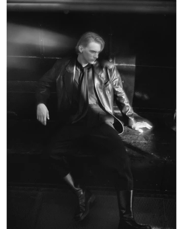 Monochrome portrait of a young man wearing a leather jacket and boots, seated against a dark background in Paris.