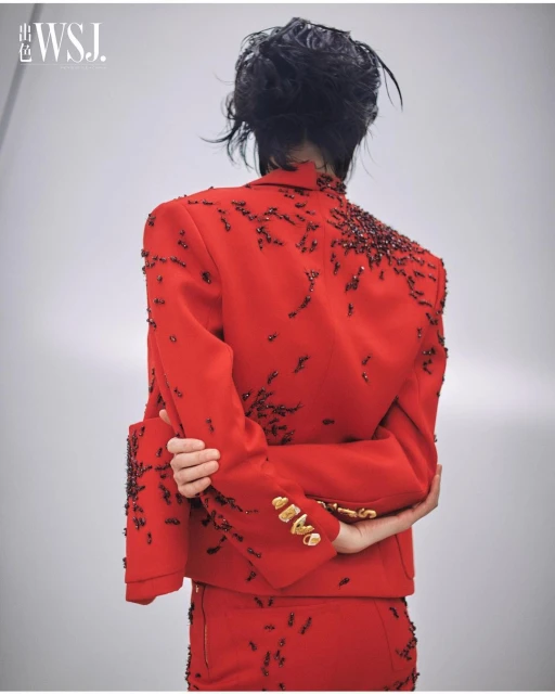 Woman in a red beaded suit, viewed from the back, standing in a light studio. Fashion editorial image featuring intricate design.