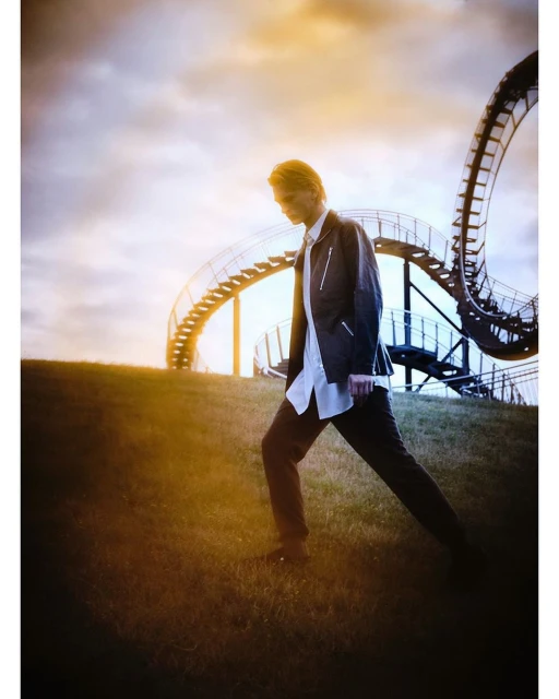 Male model poses on grassy hill beneath dusk sky and architectural sculpture. Wearing dark jacket, light shirt, dark pants. 