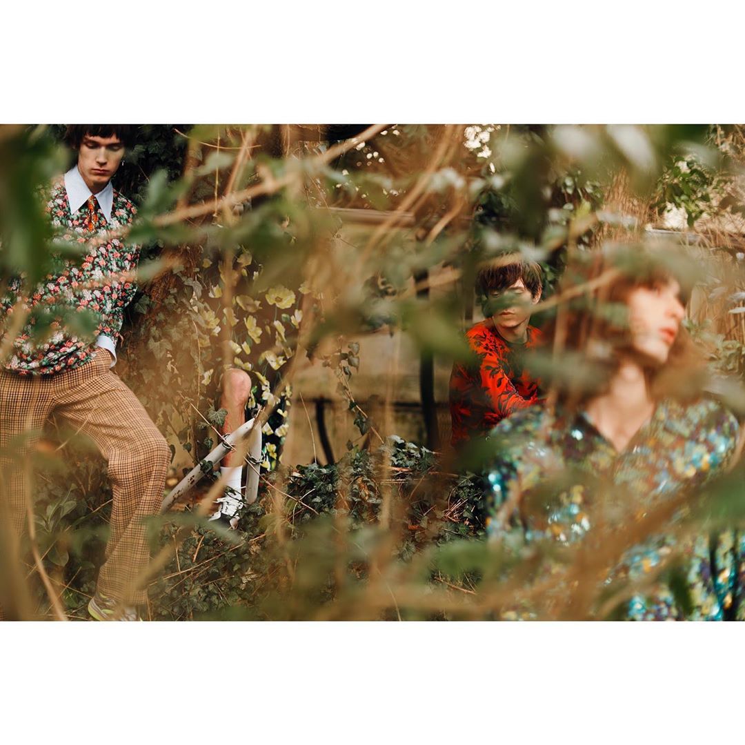 Three people pose outdoors amongst greenery in a fashion editorial image; one wears a floral shirt with plaid pants, another a glitter top.