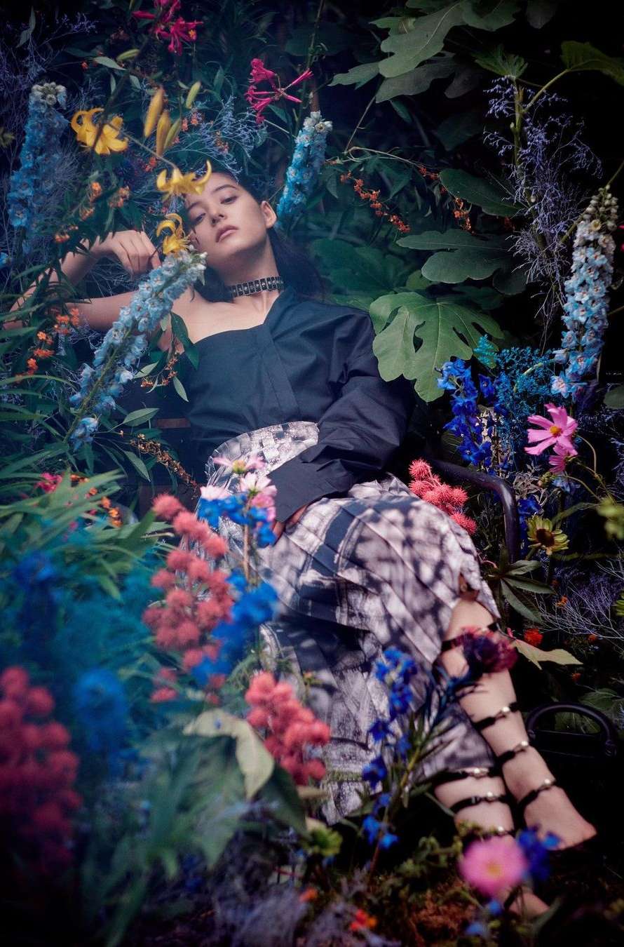 Woman in dark asymmetrical top and patterned skirt surrounded by dense garden flowers by Serge Leblon
