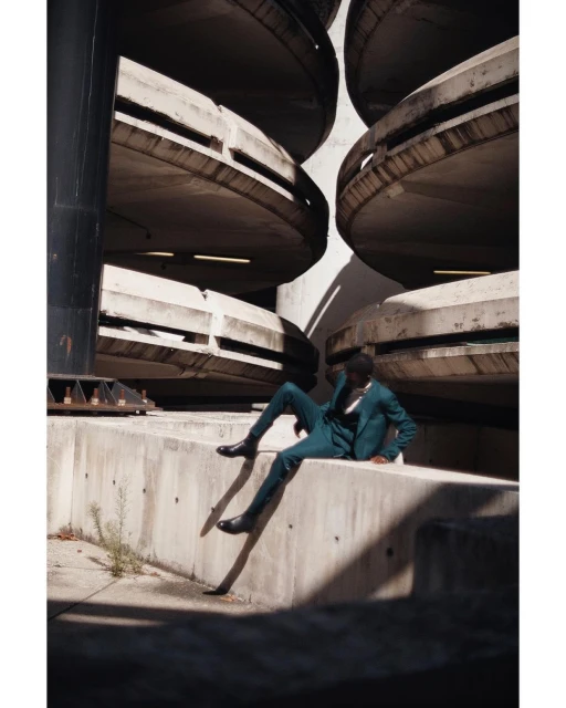 An African-American male model sits on a concrete ledge in a parkade wearing a dark teal suit, creating a stylish, urban fashion image.