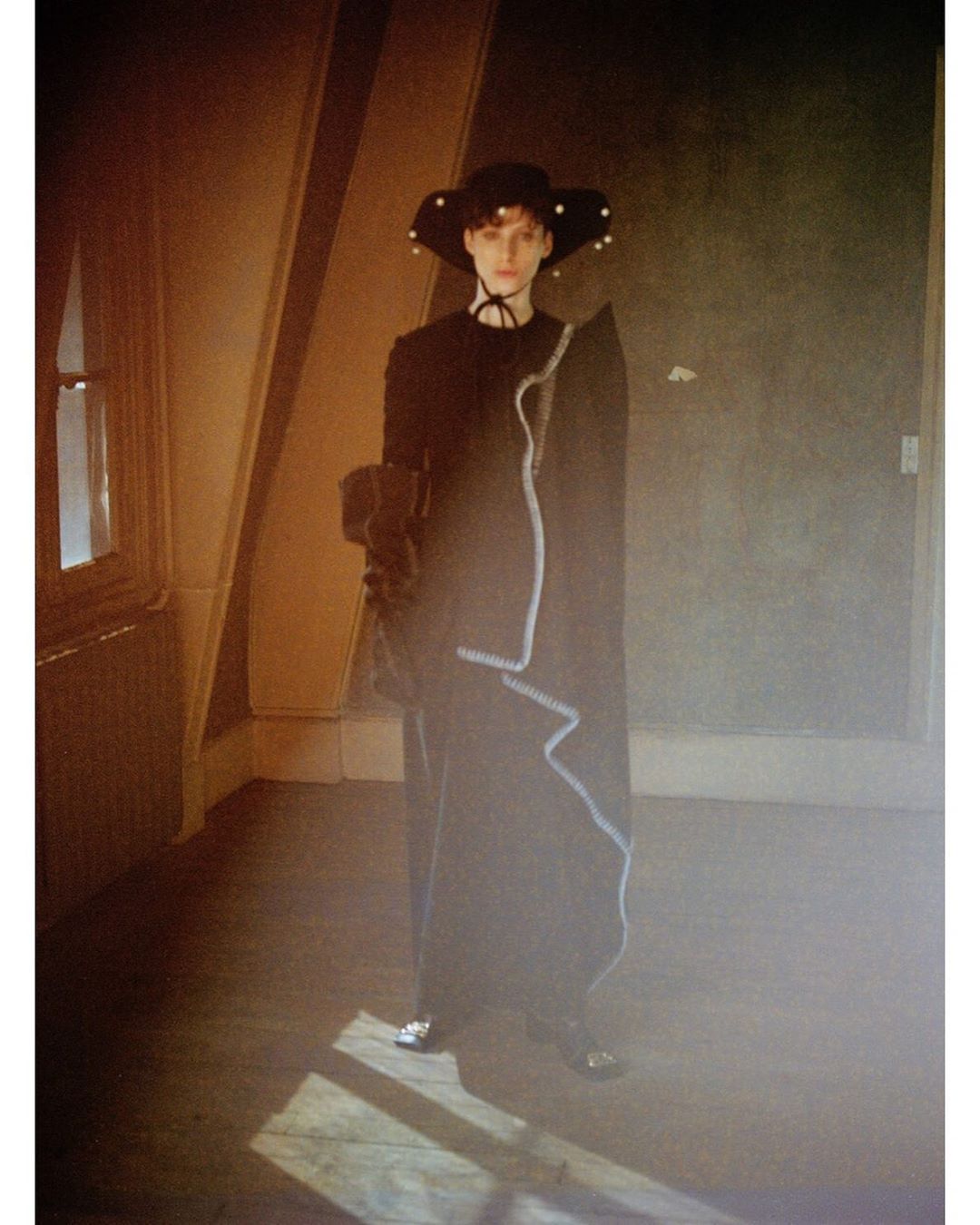 A fashion photo shows a person wearing a black dress and hat in an old room with muted lighting, showcasing editorial style.