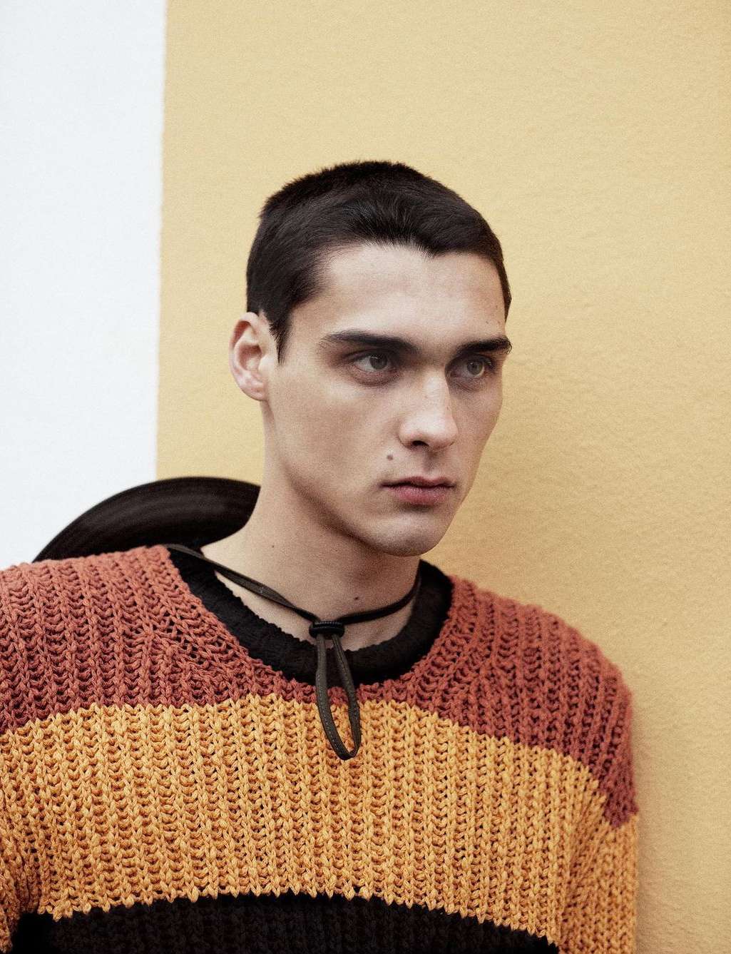 Close-up of a young man in a striped sweater against a two-tone background, looking away from the camera with a neutral expression.