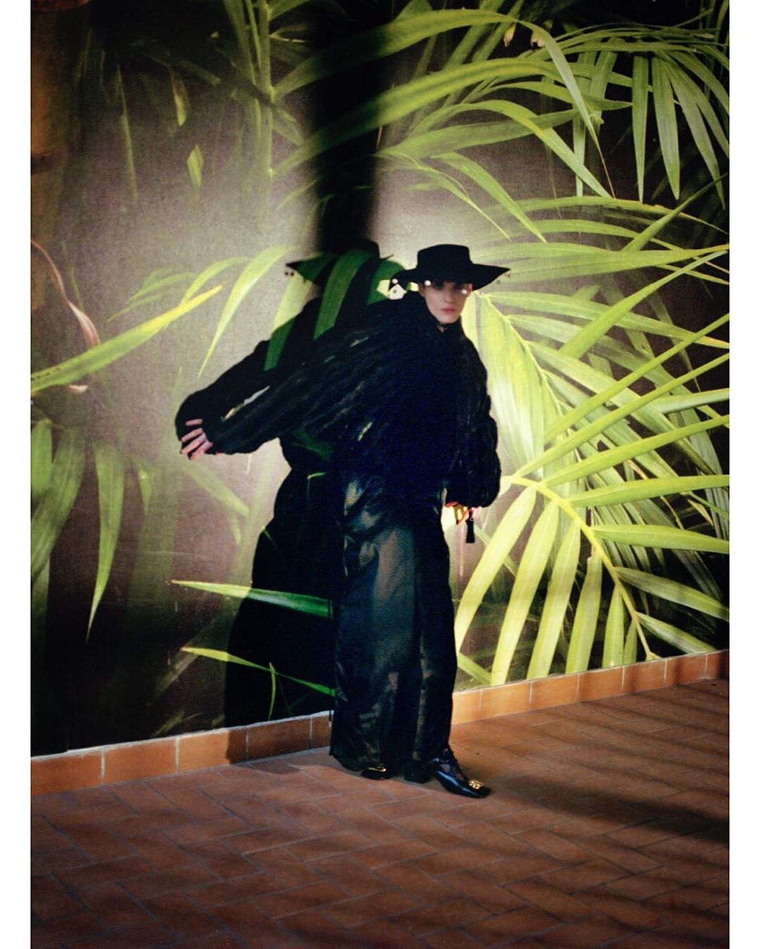 Fashion model in a black hat, black jacket, and shiny black pants poses against a backdrop of green palm fronds and a brick base.