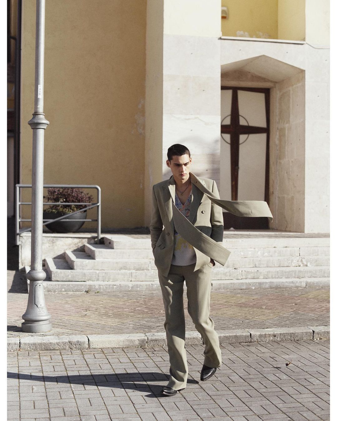 A young adult male in a beige suit walks on a bricked urban street near a light pole and the entrance to a church with a cross.