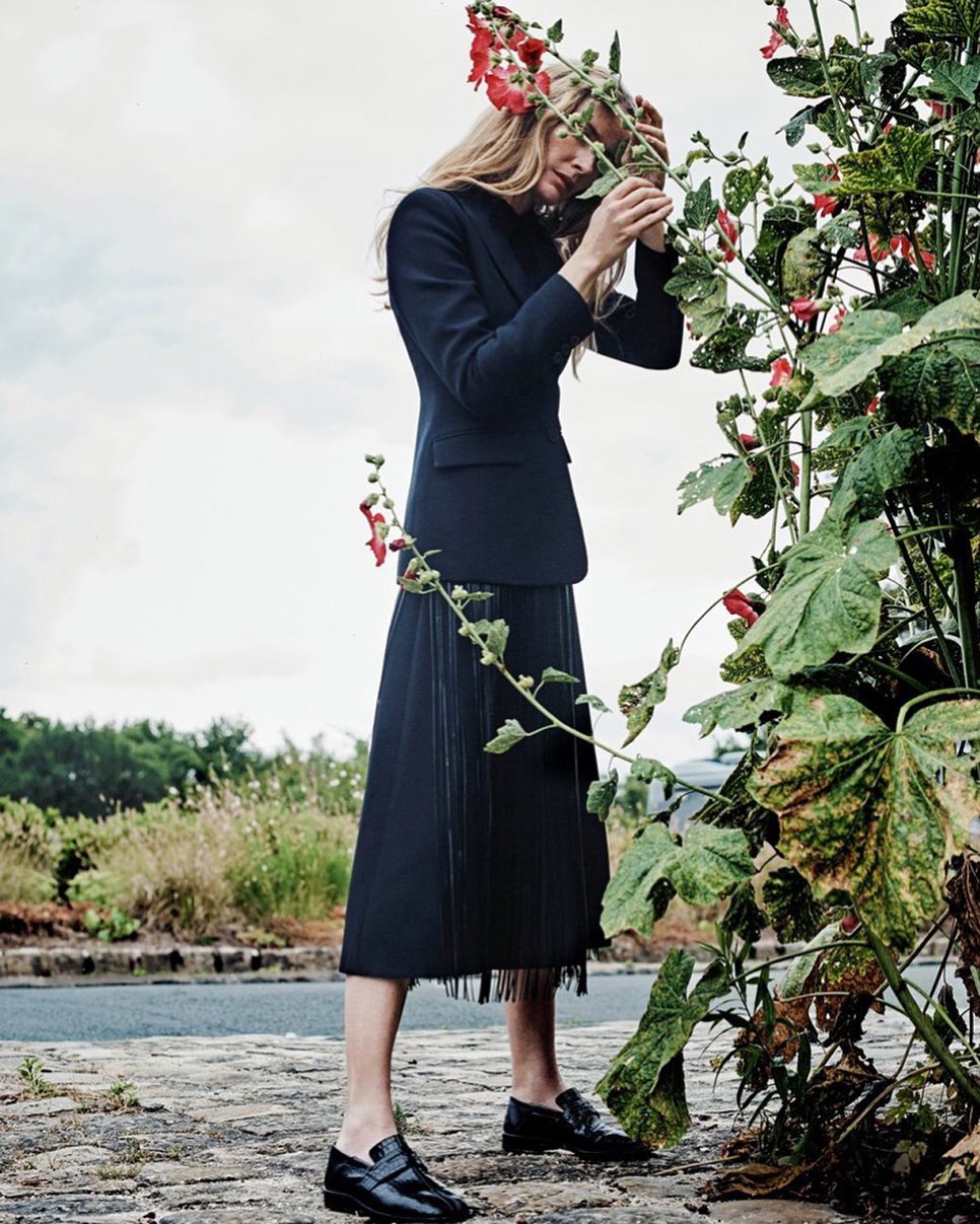 A woman in a dark blazer and skirt stands outdoors, interacting with tall flowers with red blooms, in a fashion or editorial setting.