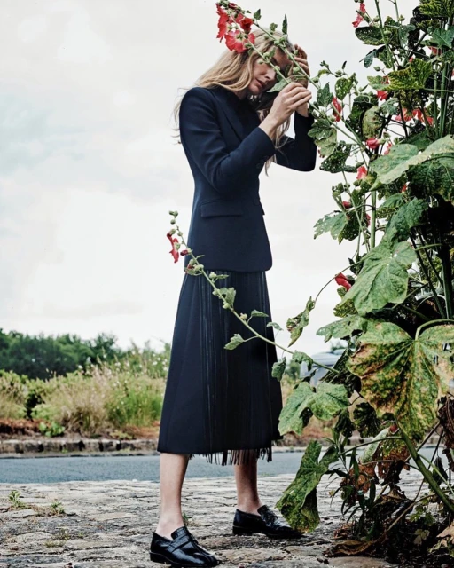 A woman in a dark blazer and skirt stands outdoors, interacting with tall flowers with red blooms, in a fashion or editorial setting.