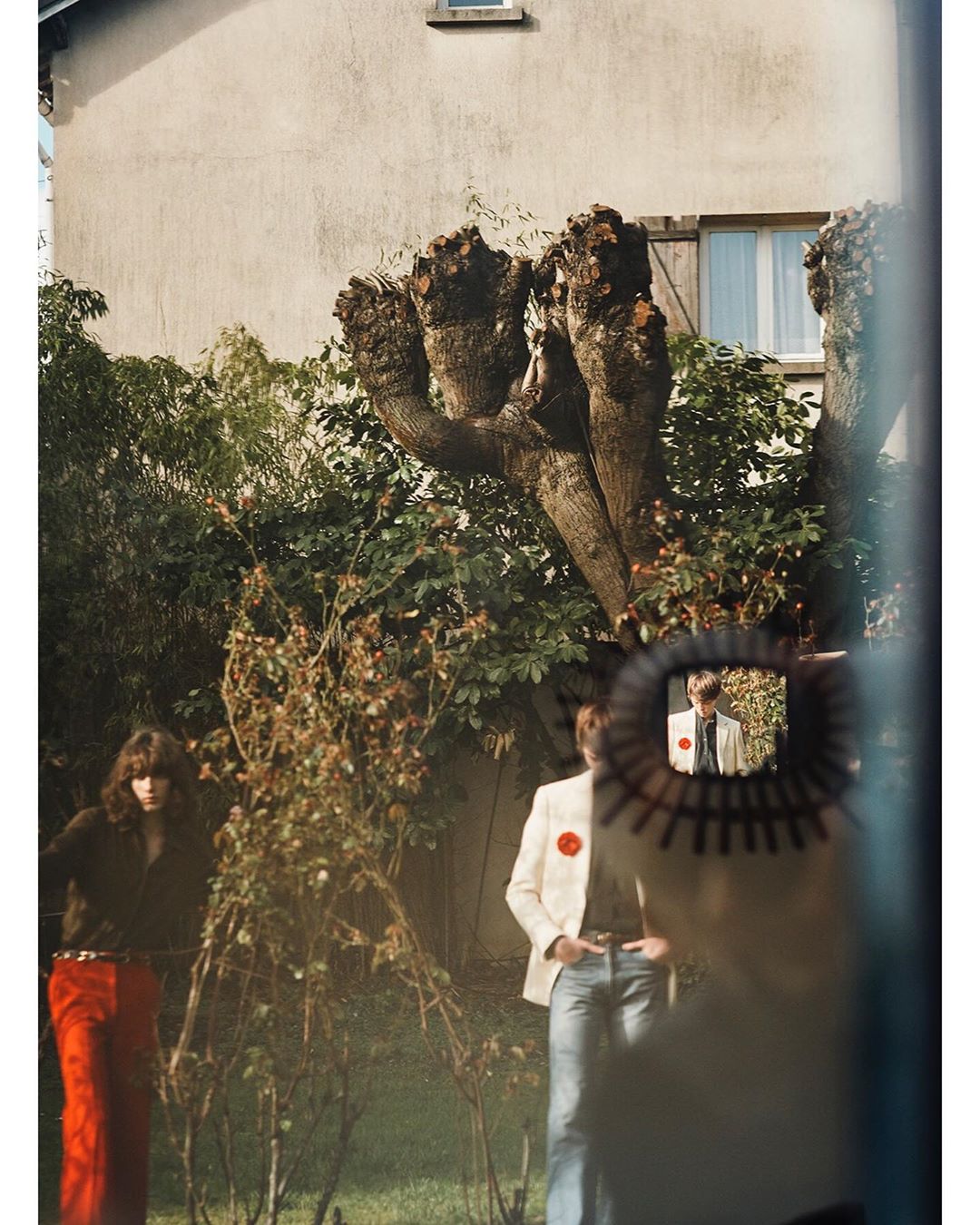 Two people in a garden setting are reflected in a small, circular window. One wears a white suit; the other has red pants and dark hair.